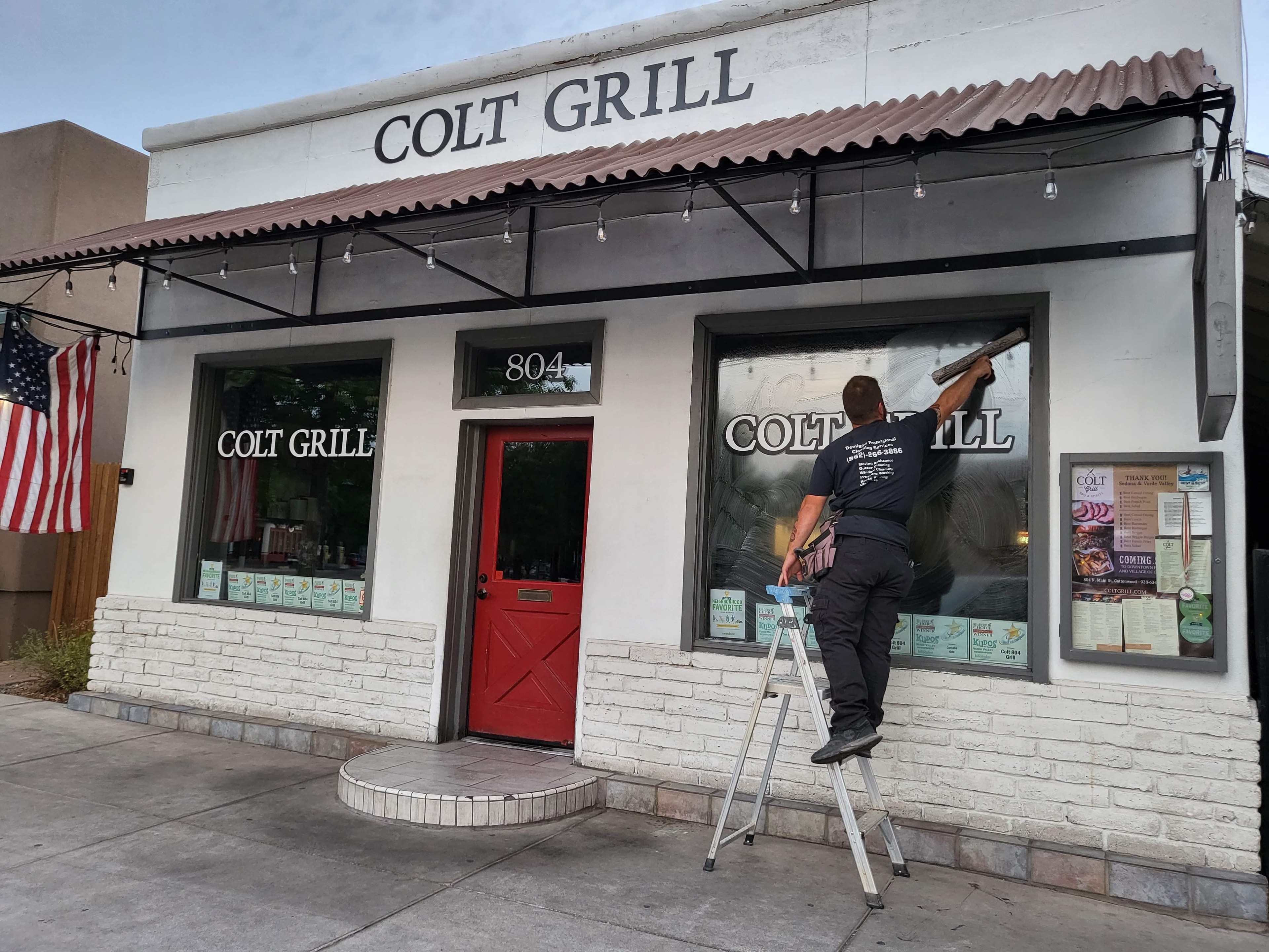 Demigod technician on ladder cleaning storefront windows at Colt Grill restaurant in Cottonwood, AZ