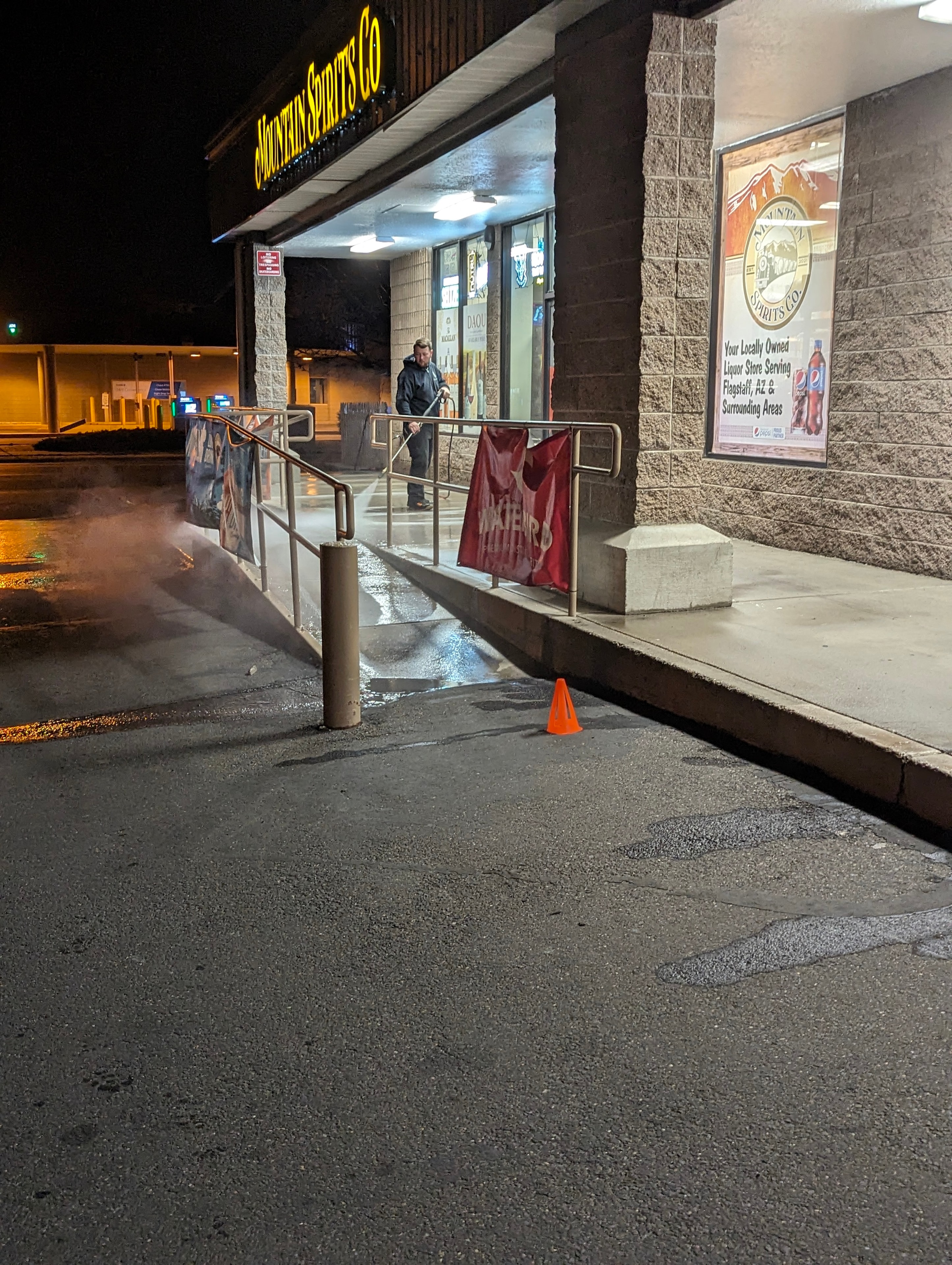 Demigod technician pressure washing the entrance and sidewalk at Mountain Spirits Co. liquor store in Flagstaff, AZ at night with steam rising