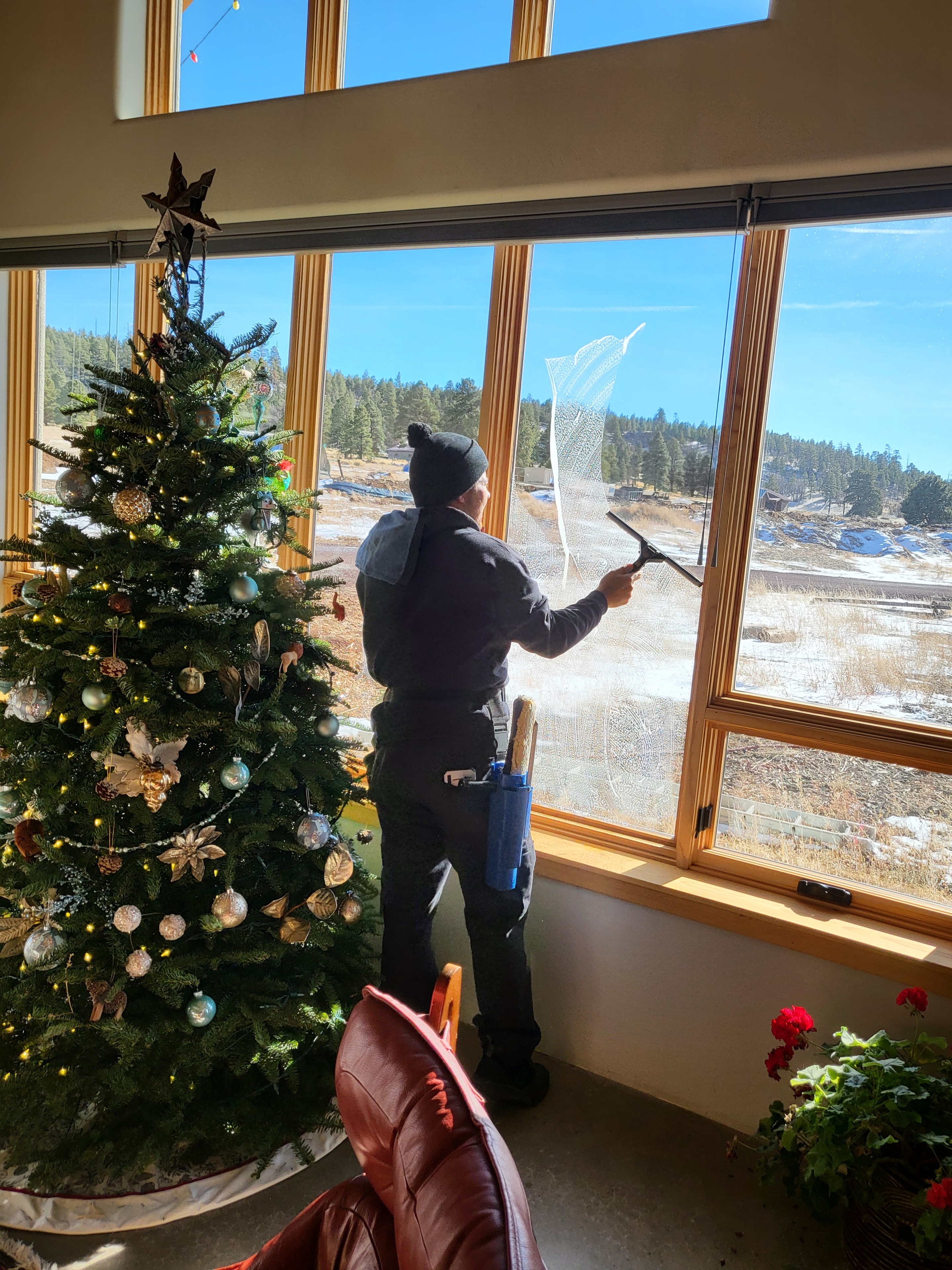 Demigod technician squeegee cleaning large interior windows at a Flagstaff home with snow-dusted pines visible outside and Christmas tree in foreground