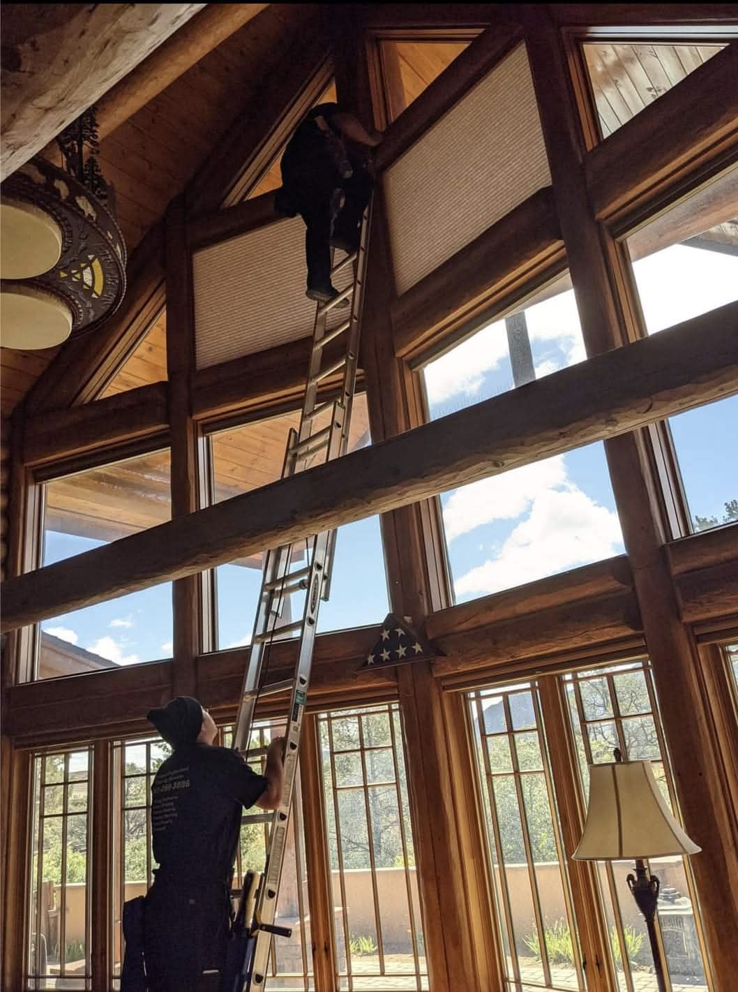 Two Demigod technicians on extension ladders cleaning floor-to-ceiling windows inside a Flagstaff log cabin with exposed timber beams