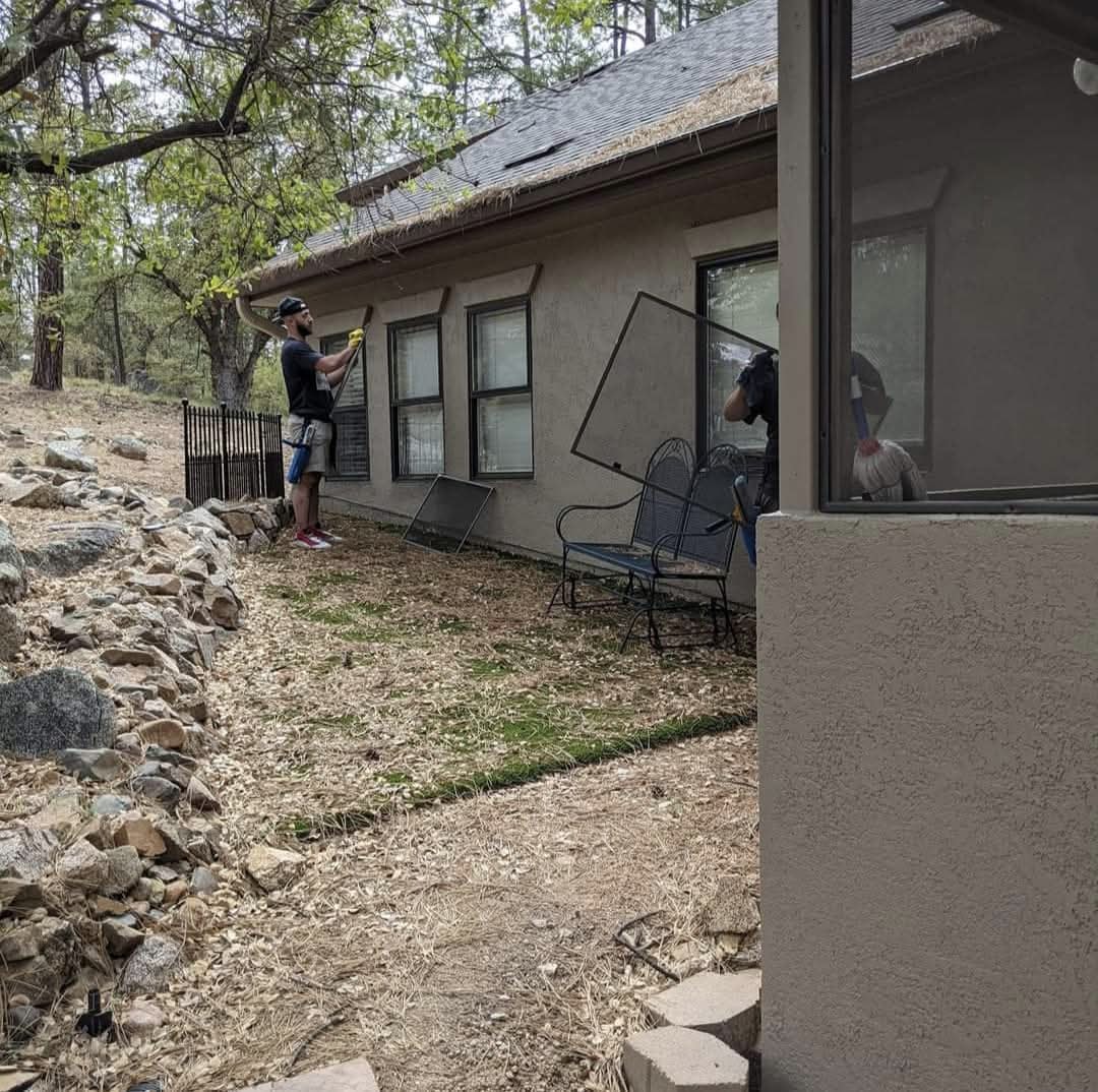 Two Demigod technicians cleaning windows on a stucco home exterior in Flagstaff, AZ surrounded by ponderosa pines and rock landscaping