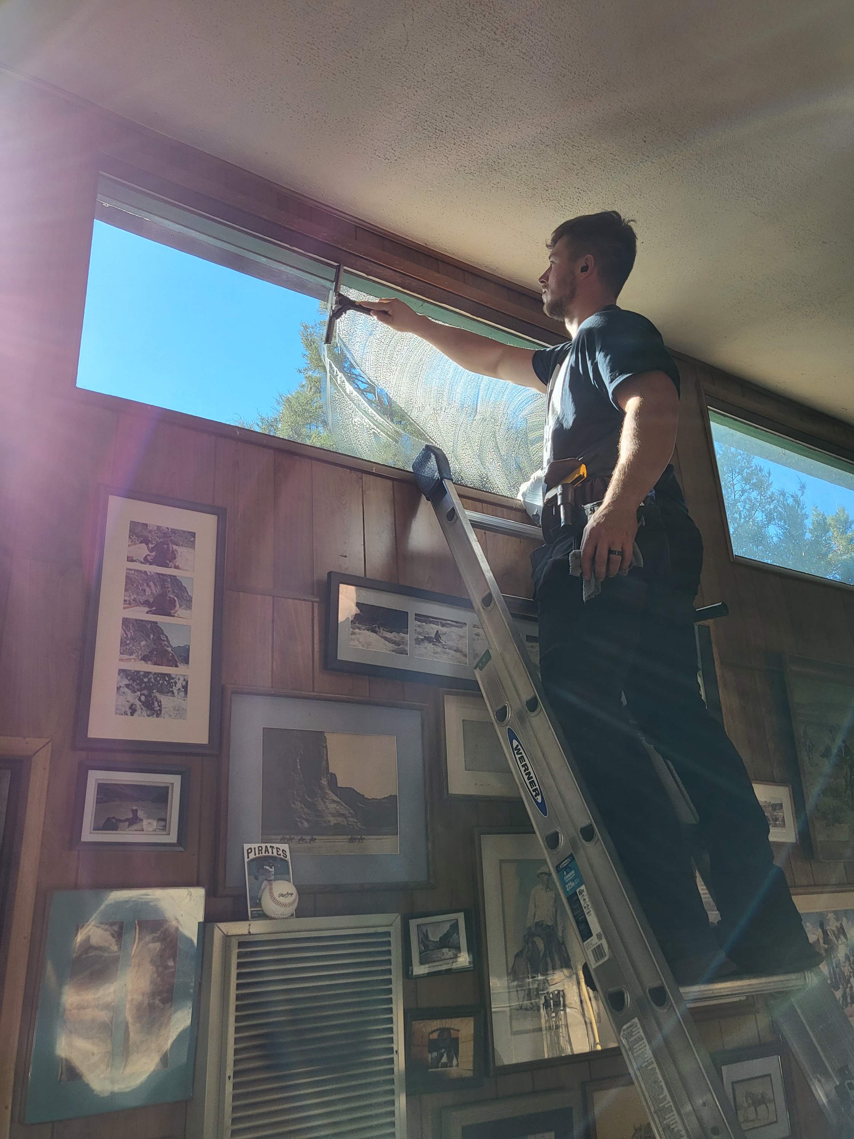 Demigod technician on ladder cleaning interior windows at a Prescott residential home with art-filled walls