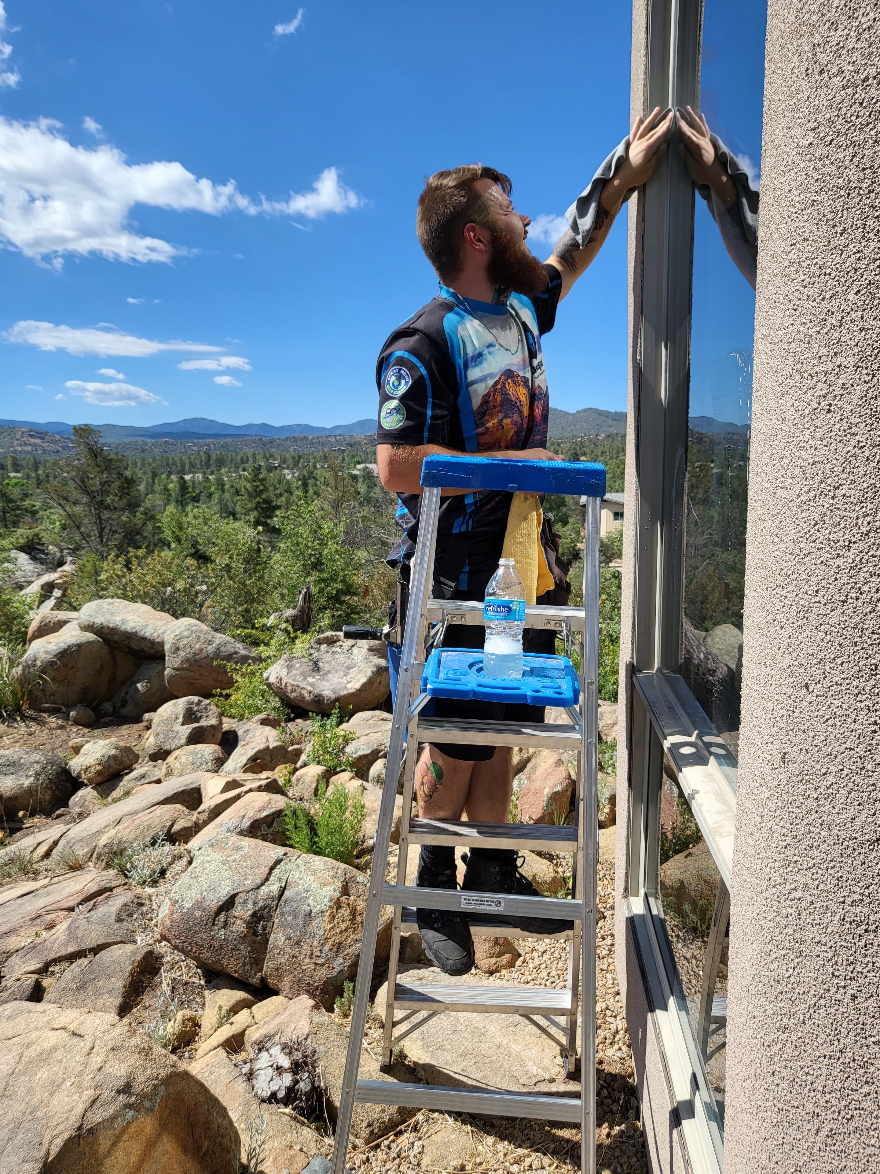 Demigod technician on ladder cleaning windows at a Prescott hillside home built on granite boulders with mountain views