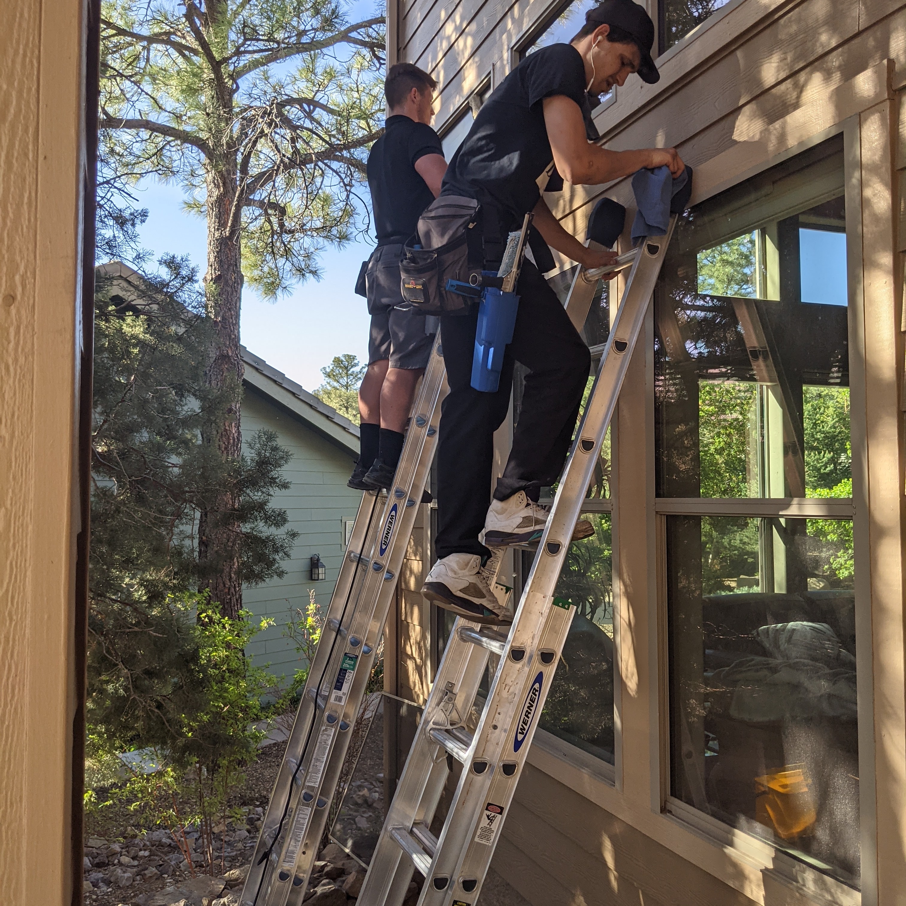 Two-person Demigod window cleaning crew on extension ladders cleaning windows on a Prescott home surrounded by ponderosa pine trees