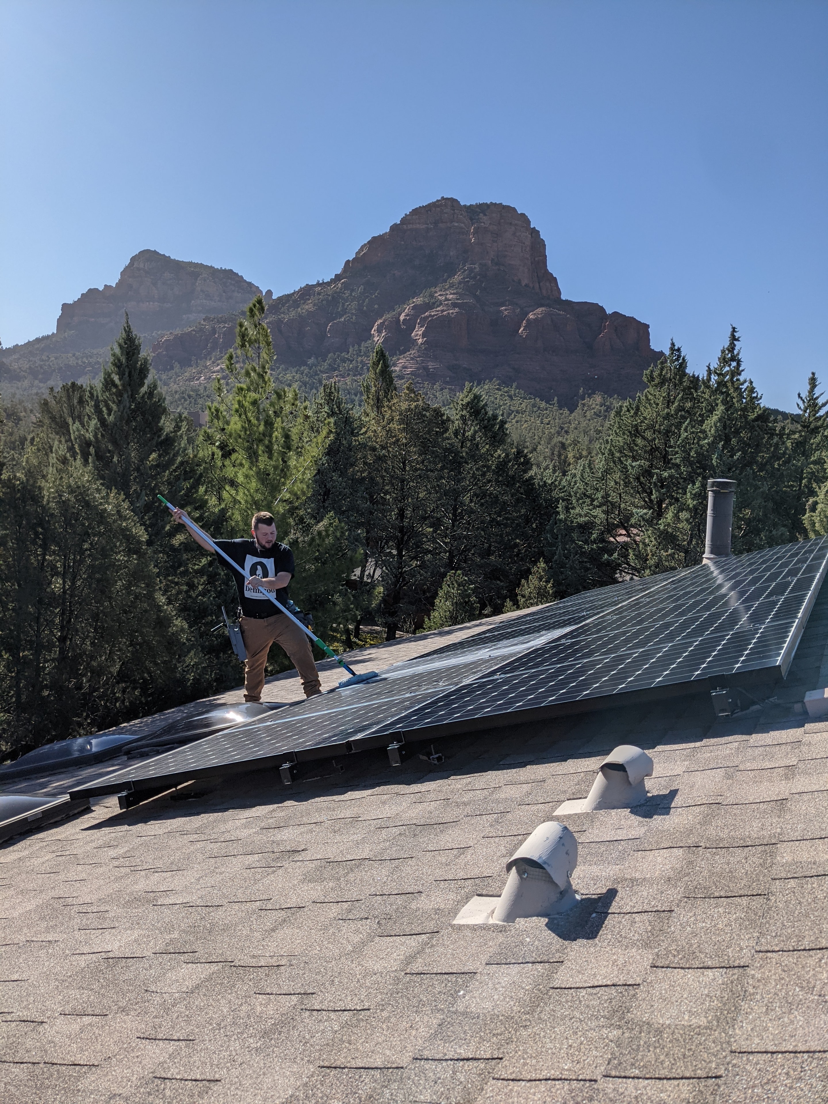 Demigod technician cleaning solar panels on a shingle roof in Sedona with red rock mountain backdrop