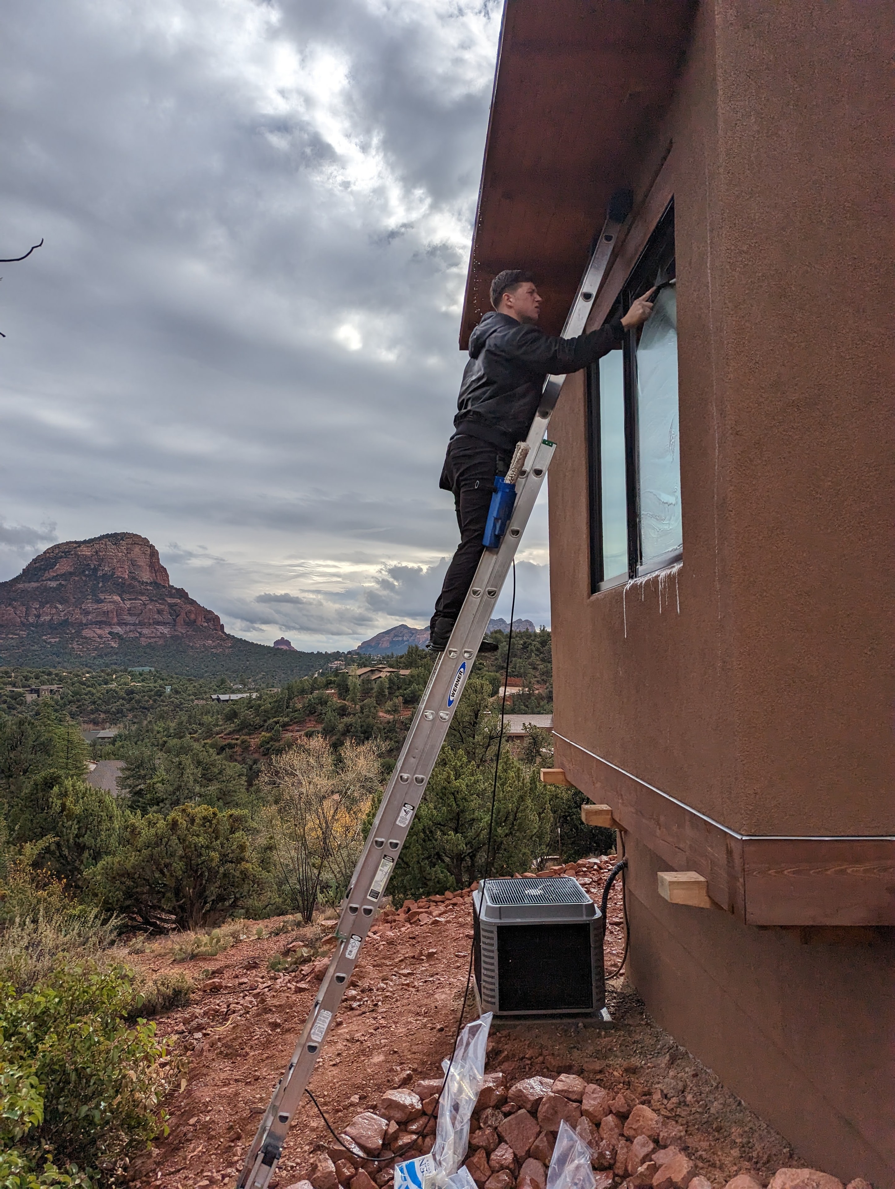 Demigod technician on extension ladder cleaning high windows on a stucco Sedona home with red rocks in background